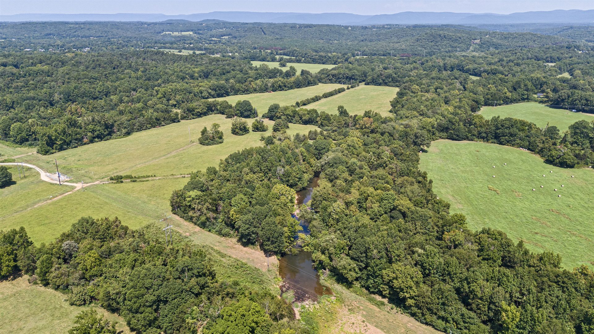 Aerial view showing open fields, wooded corridors, a visible road and creek in the surrounding landscape